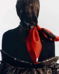 Back view of a person in a black puffer jacket with a red and black patterned scarf tied at the neck against white background