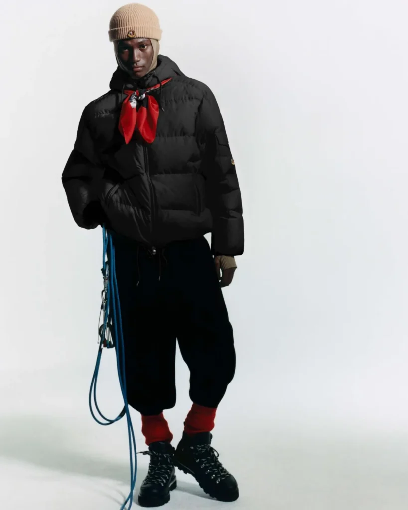 Model in a black puffer jacket with a red scarf, beige knit cap, black pants and hiking boots, against a white backdrop