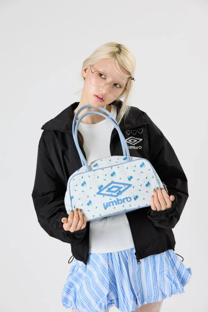 Blonde model in glasses holds a white floral Umbro handbag, wearing a black jacket and blue gingham skirt against a pale