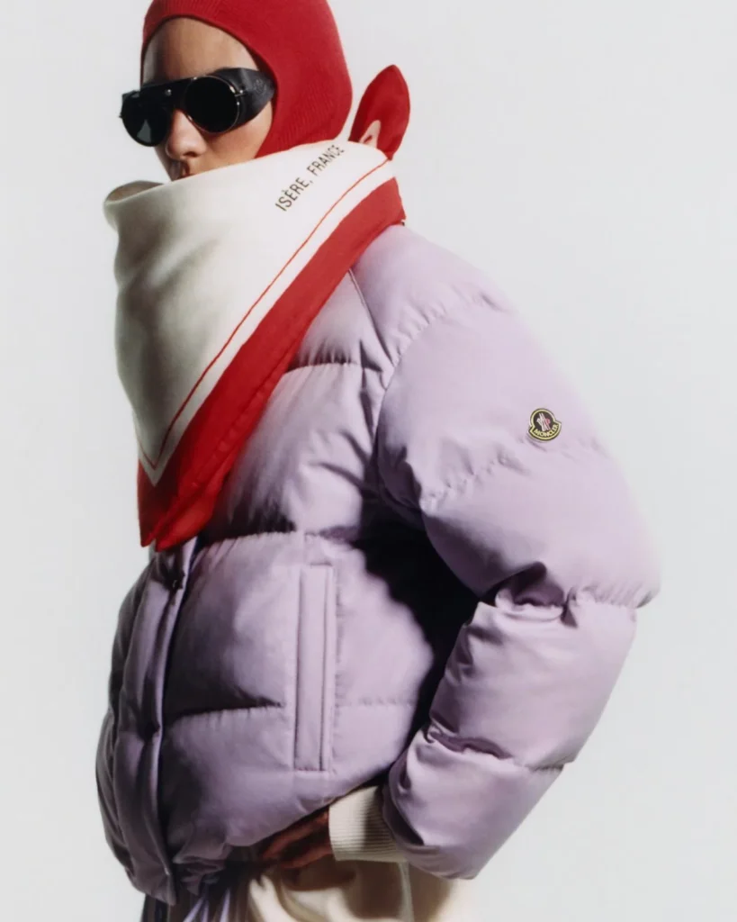 Person in a lavender puffer jacket with a red hood and scarf, black sunglasses, and Moncler badge against a white background