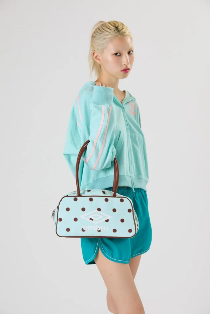 Model in a mint zip-up hoodie and teal shorts holds a polka-dot Umbro duffel bag against a white background