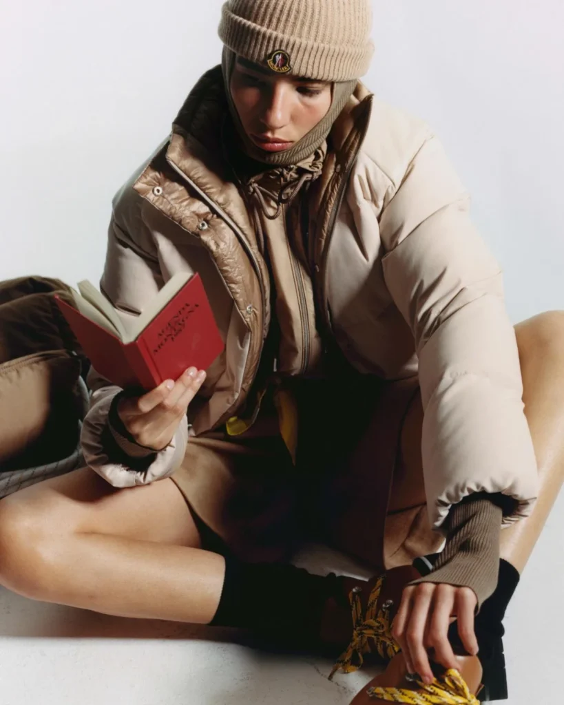 Person in a beige puffer jacket and ribbed beanie reads a red book while seated on the floor beside a tan chair
