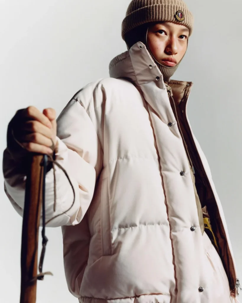 Person in a white puffer jacket and brown beanie holds a wooden ski against a plain light background