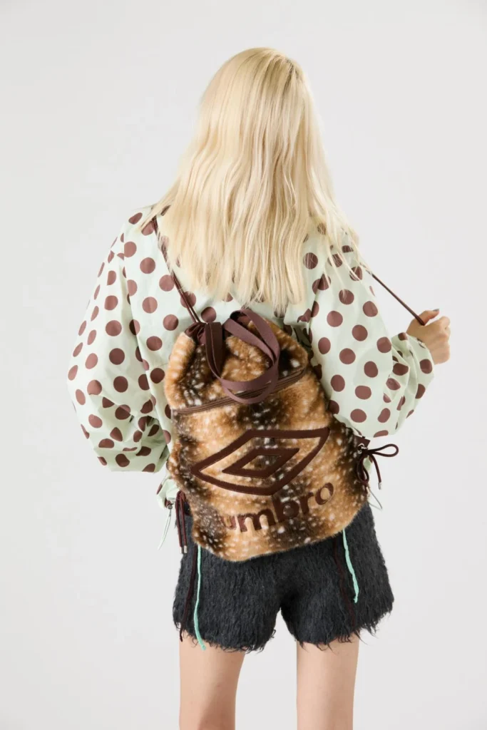 Blonde model seen from behind wearing a brown furry Umbro-logo backpack over a polka-dot blouse and black shorts