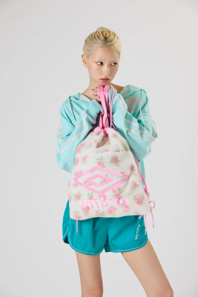 Model holds a cream floral-fleece Umbro bag with pink straps, wearing turquoise sportswear against a gray studio backdrop