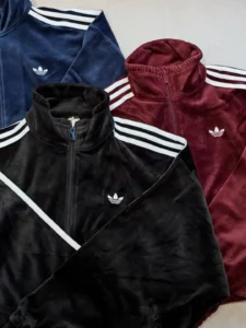 Three Adidas velvet track jackets in black, burgundy, and navy with white stripes and trefoil logos laid flat