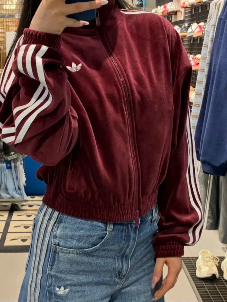 Person wearing a burgundy Adidas track jacket and blue jeans in a clothing store, face obscured by phone