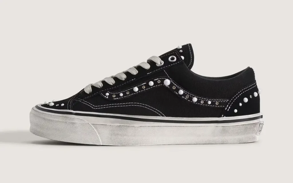 Black low-top sneaker with white laces, stitching, rubber sole, and studs along the side panels on a plain background