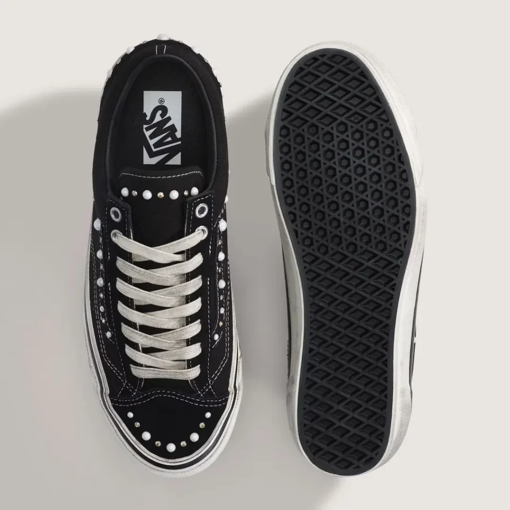 Pair of black Vans shoe, one top-down with white laces and studs, one showing a black waffle sole, on a pale background