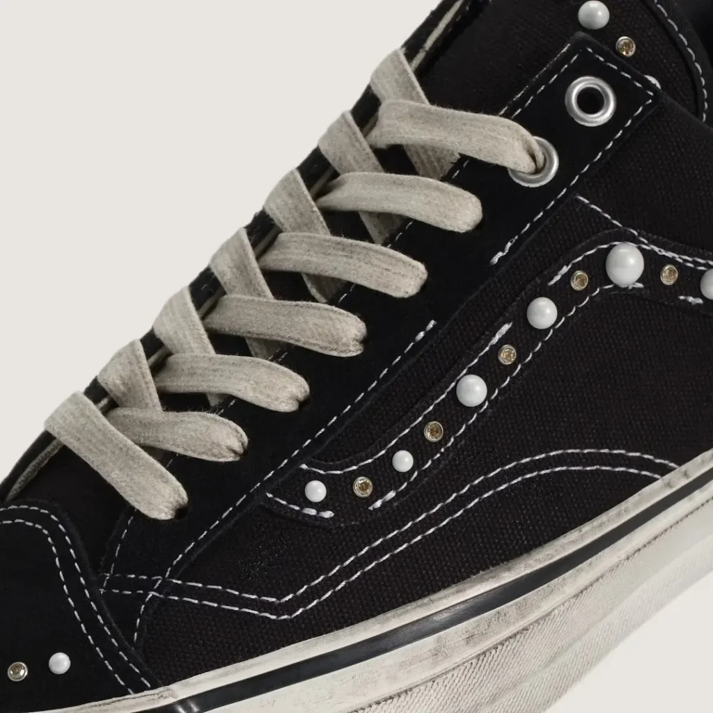 Close-up of a black high-top sneaker with beige laces, white stitching, and pearl-like studs along the side panel