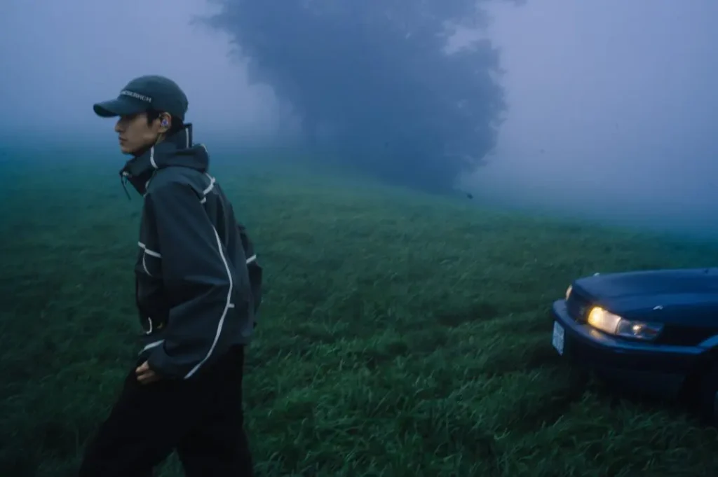 Person in a dark cap and reflective-trim jacket walks through a foggy field beside a car with headlights on