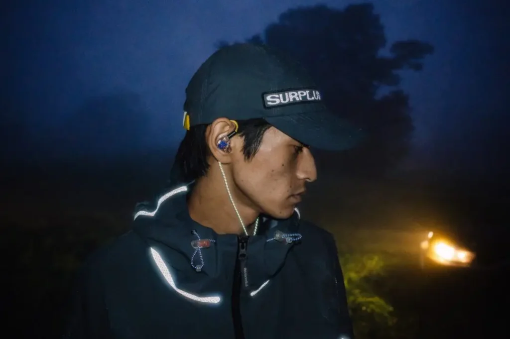 Person in a dark cap and rain jacket with reflective details, wearing an ear monitor outdoors at night
