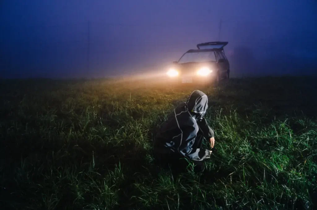 Hooded person crouches in a dark grassy field as a car with headlights on faces them through mist
