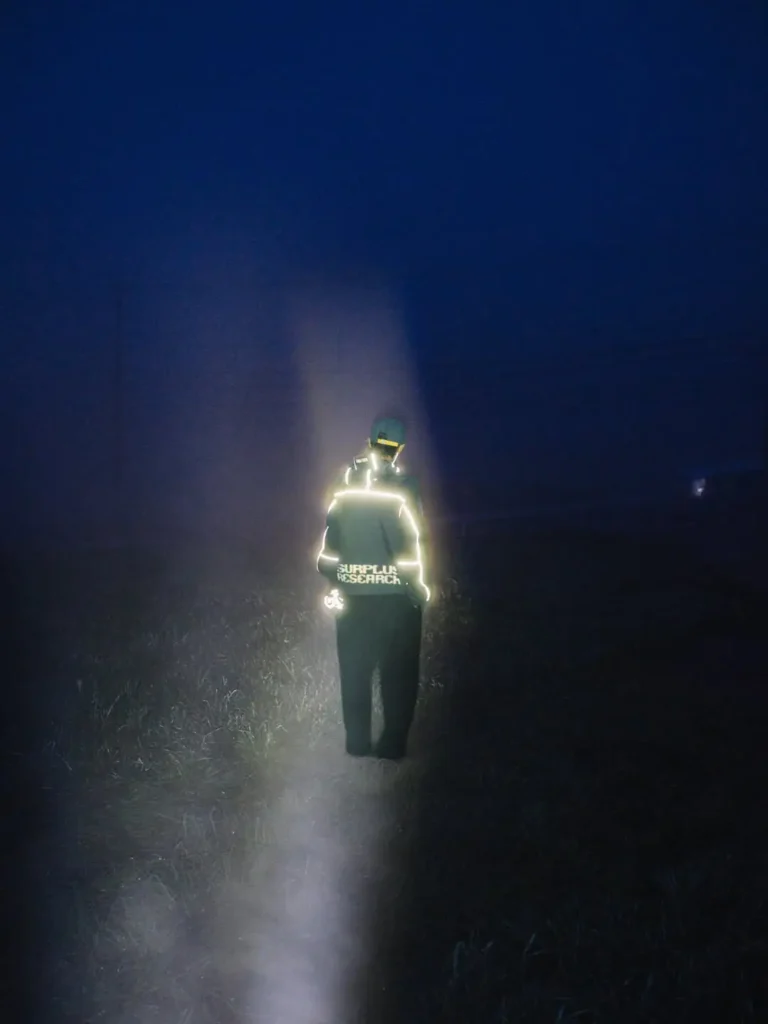 Person in dark clothing with reflective piping and “SUPPLUX RESEARCH” text stands in a flashlight beam at night