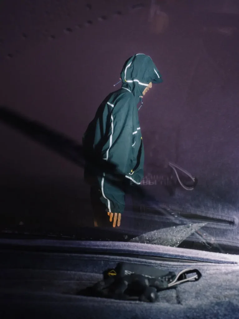 Person in a dark green hooded tracksuit with reflective stripes, seen in profile against a dark purple backdrop