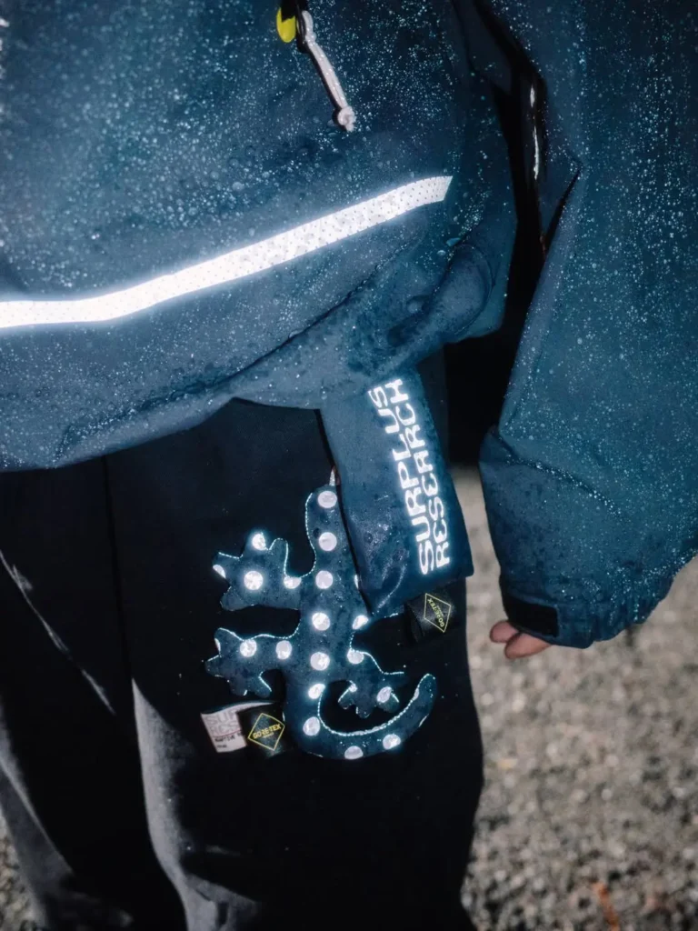 Close-up of a rain-speckled navy jacket with reflective stripe, zipper, and white Superdry Research lettering over black