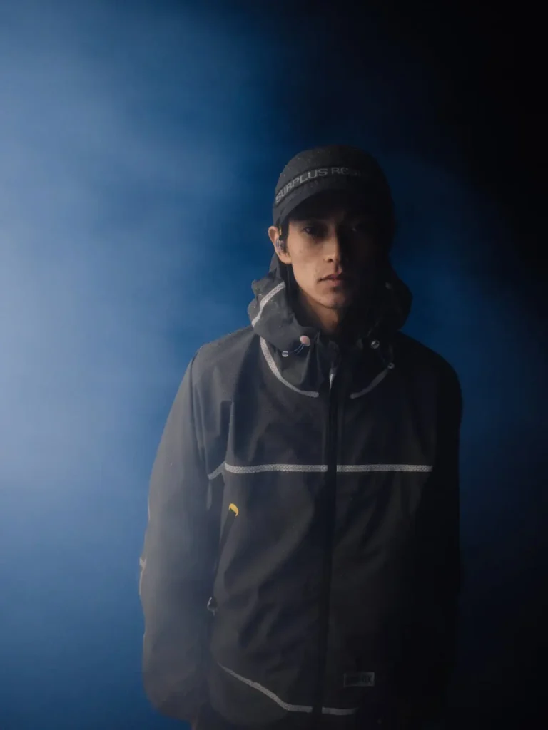Person wearing a black hooded jacket with reflective stripes and a black cap, standing against a dark blue background