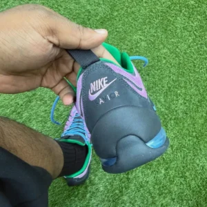 Close-up of a hand holding a purple, green, and black Nike football boot over artificial grass