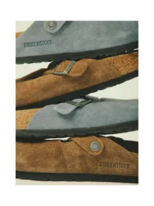 Stacked close-up of gray and brown Birkenstock suede sandals with buckles and black soles against a pale background