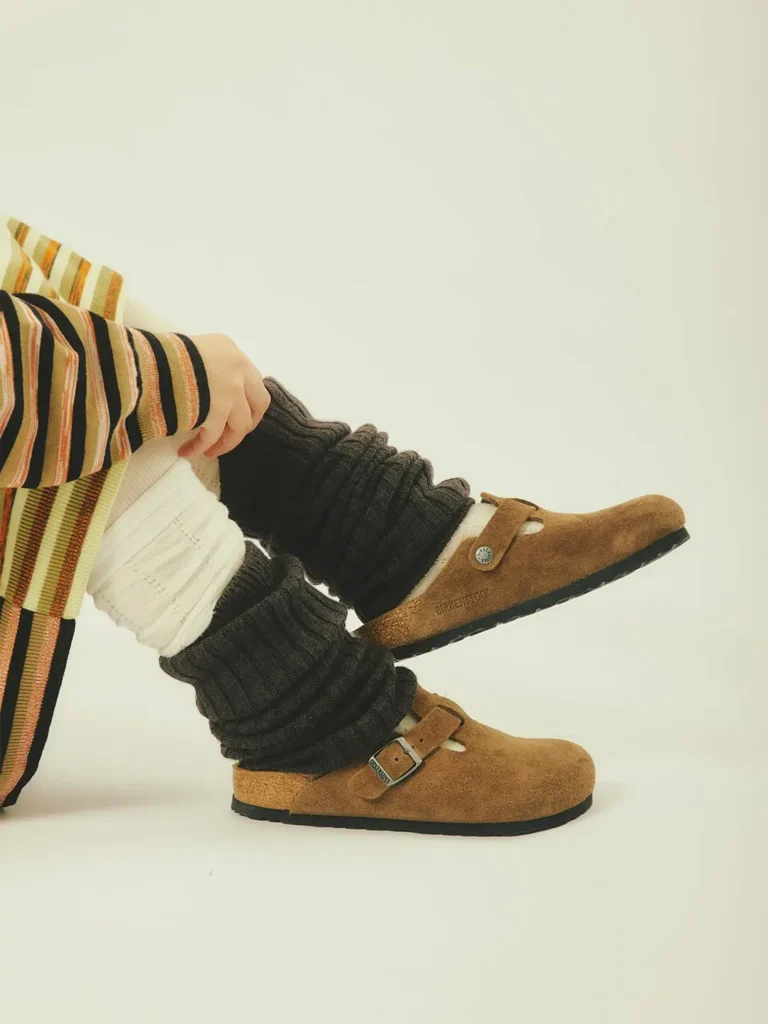 Person wearing striped pants, cream socks, and brown buckled shoes with black leg warmers reclining against a pale background