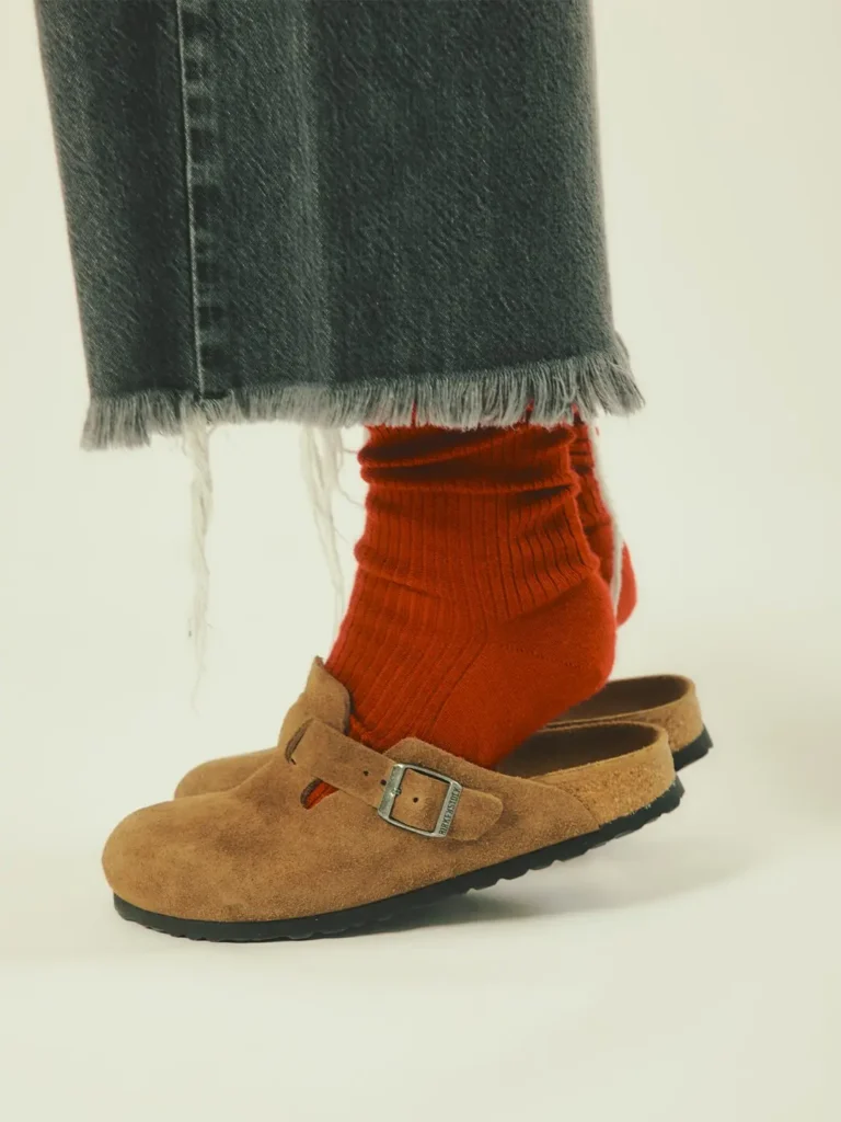 Close-up of a foot in a rust-red sock and tan suede buckle clog beneath a frayed dark denim hem
