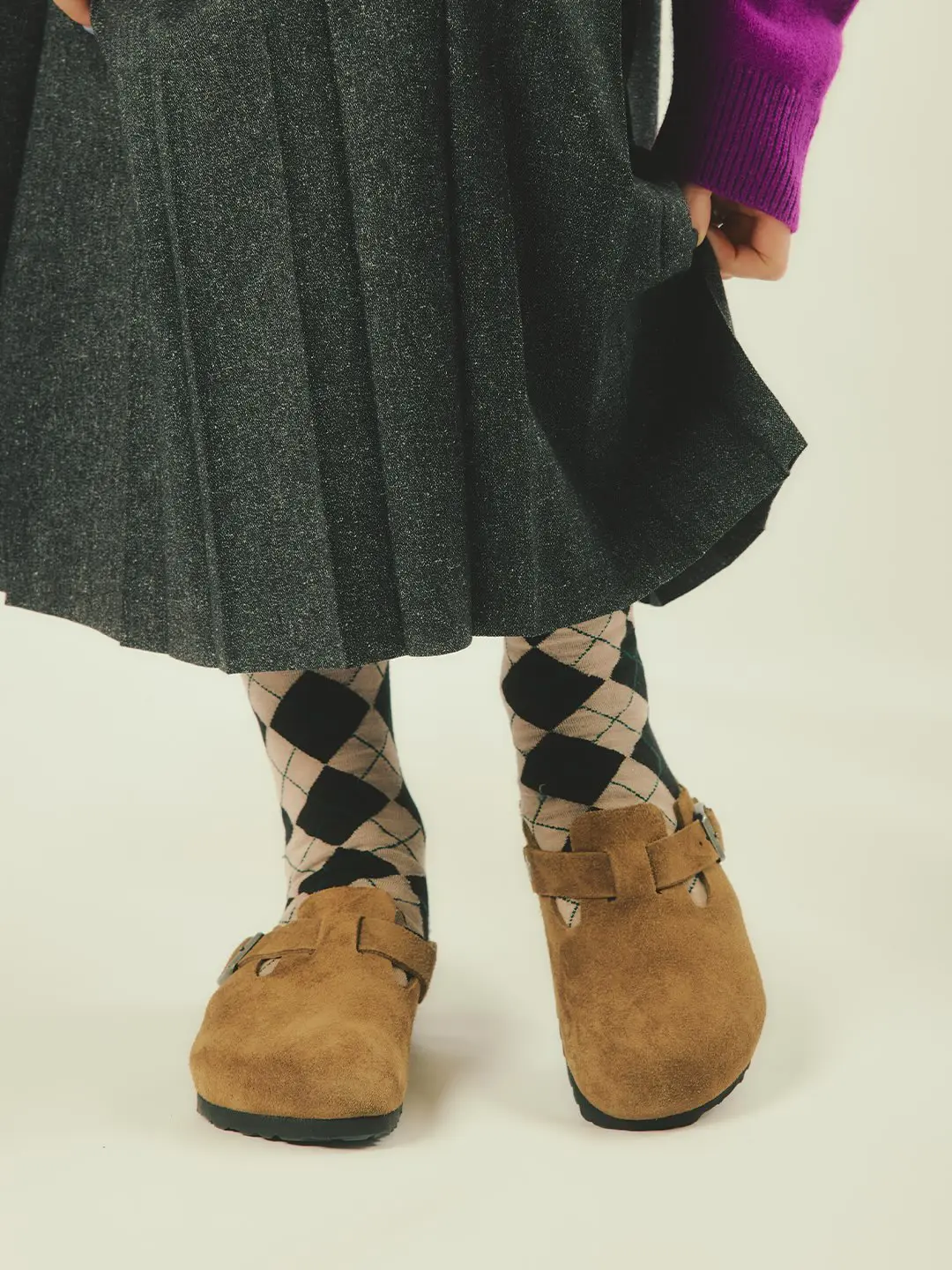 Cropped figure wearing a charcoal pleated skirt, purple sweater, argyle socks, and tan suede buckle clogs against a pale