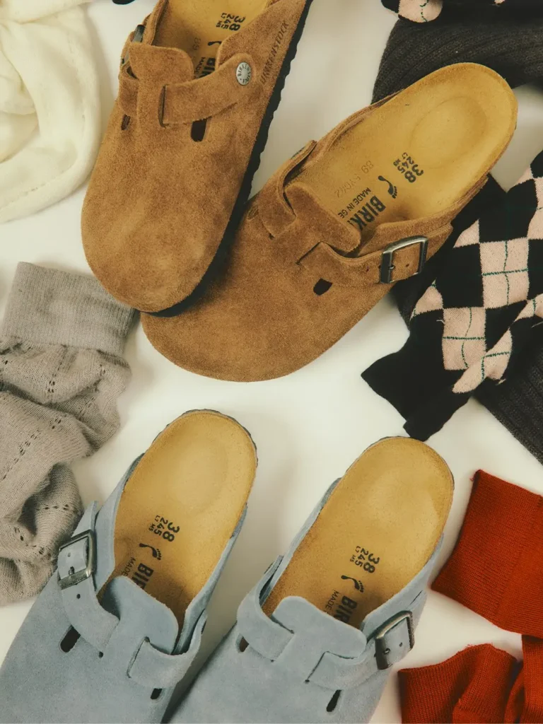 Tan and light gray suede buckle clogs arranged with folded cream, gray, black-and-pink, and orange knitwear