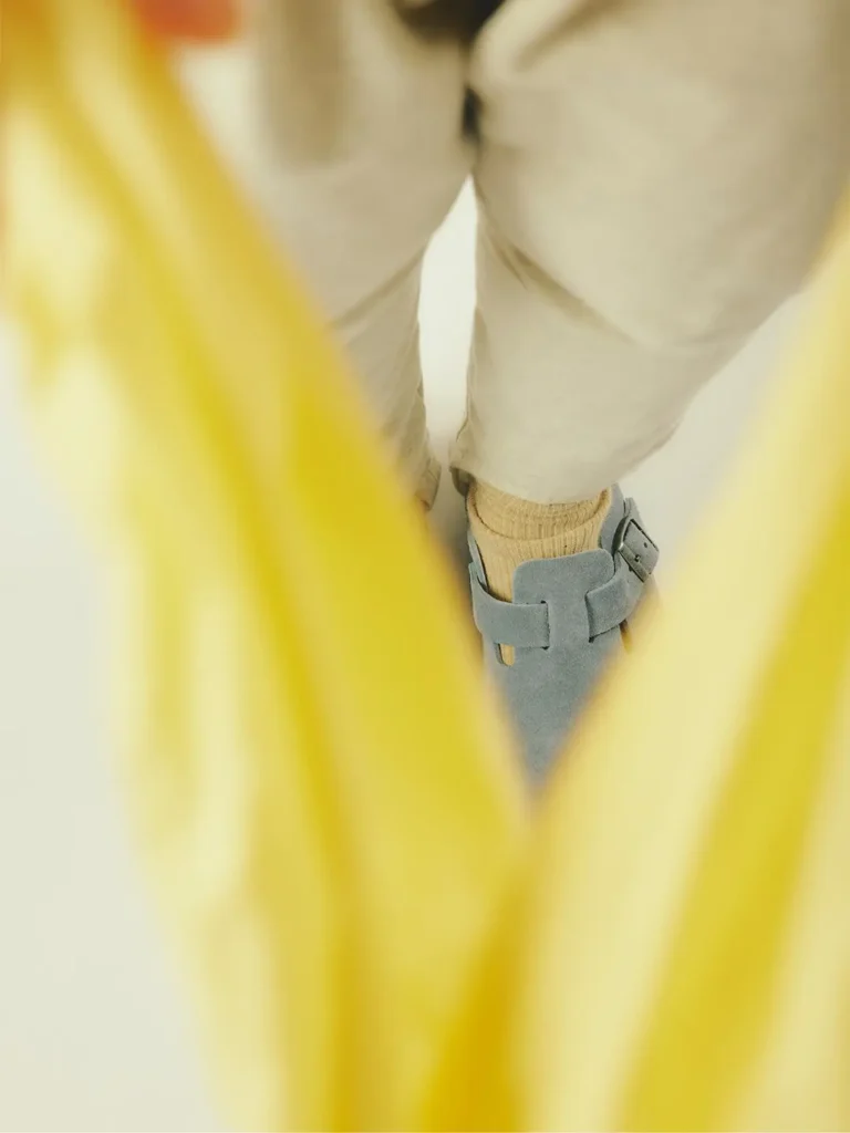 Close-up of cream trousers, beige socks and gray Mary Jane shoes, partly obscured by a bright yellow garment