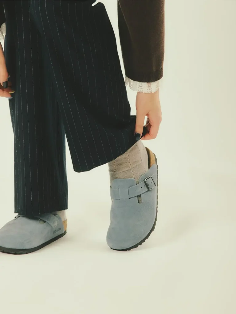 Cropped figure wearing a black pinstriped skirt and jacket with lace cuffs, beige socks, and gray buckled clogs against a