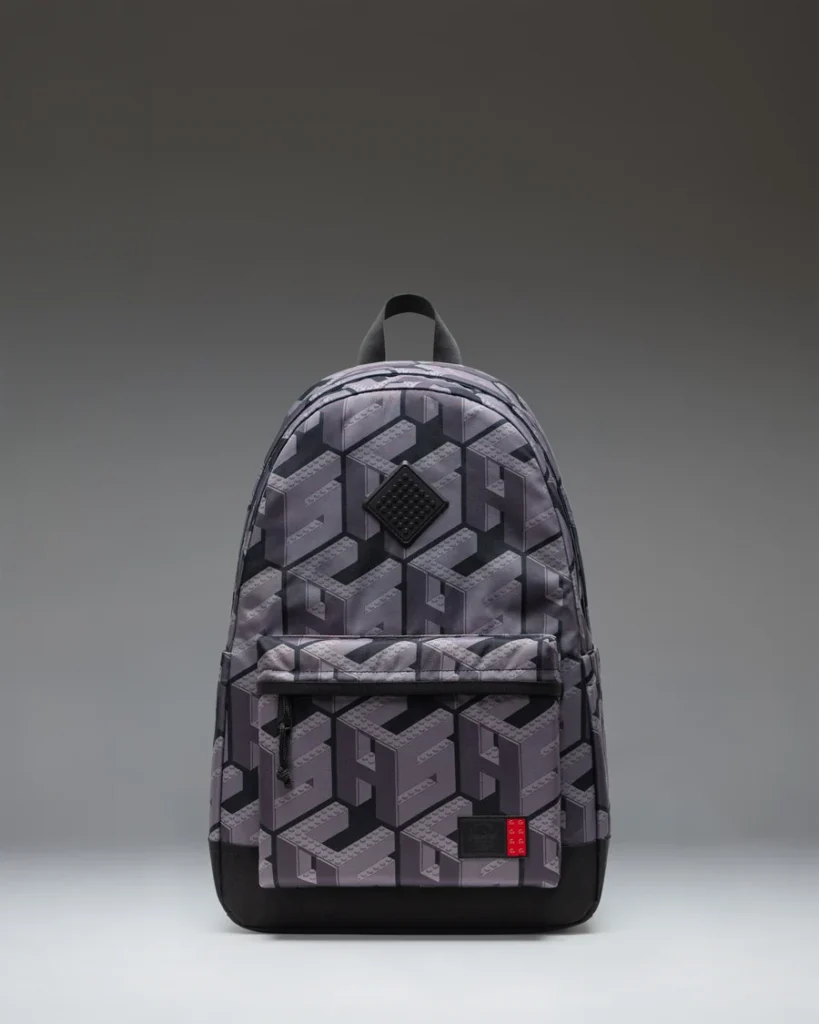 Gray and black geometric-patterned backpack with a front zip pocket and small red label, centered against a gray backdrop