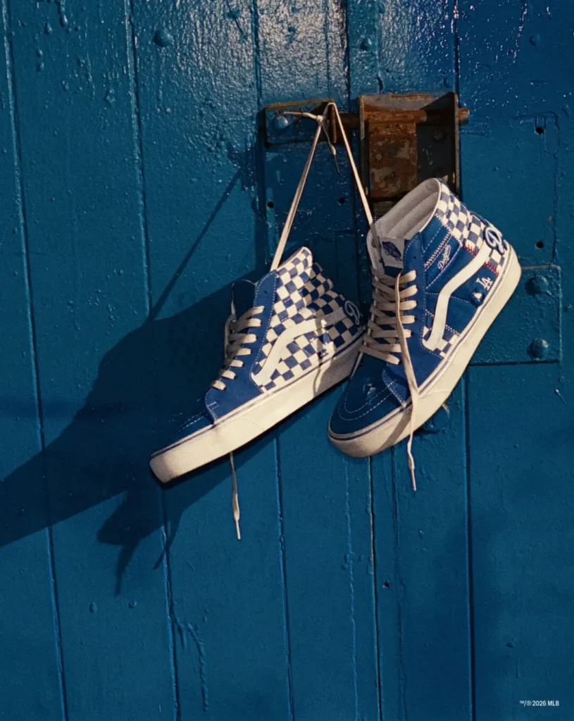 Blue-and-white high-top sneakers hang by their laces on a blue wooden wall; one has a checkerboard pattern and LA logo