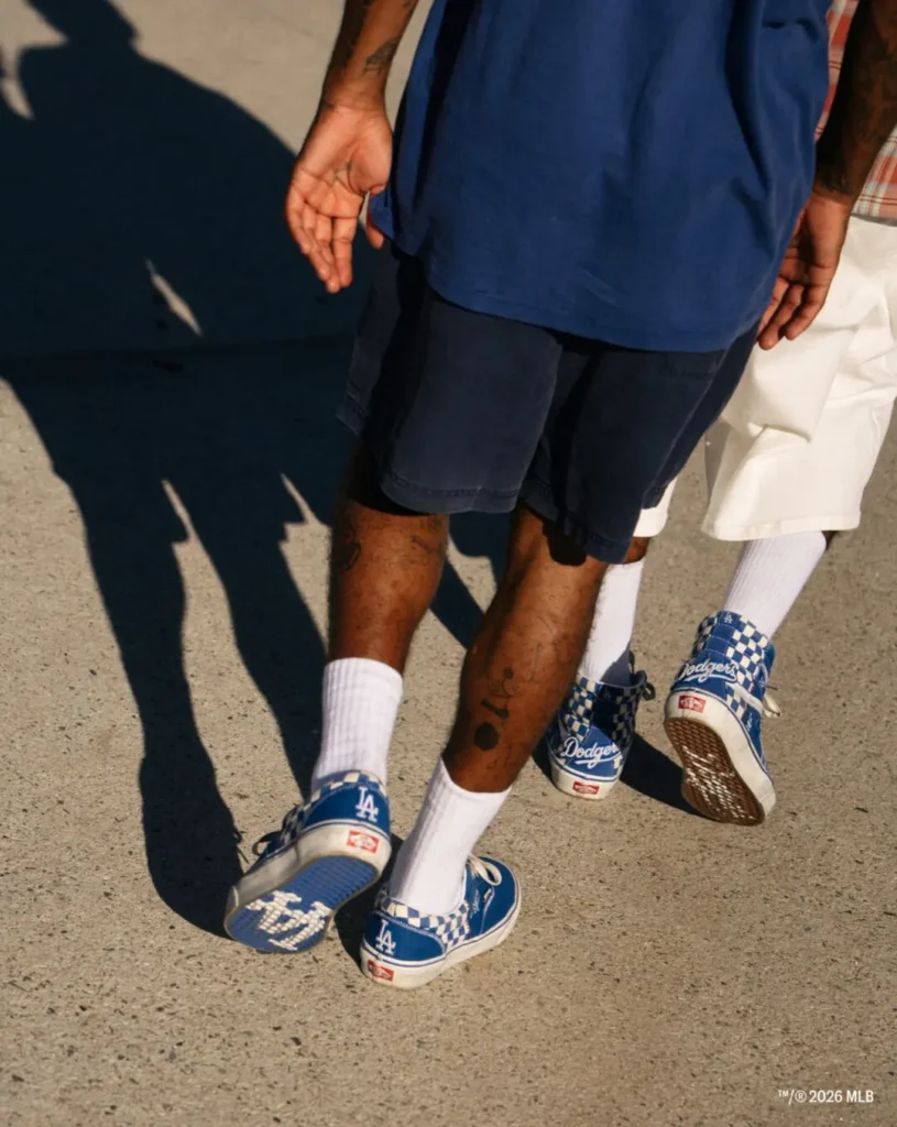 Two people walking on pavement in blue Dodgers sneakers with checkerboard panels, white socks, and shorts; long shadows cast