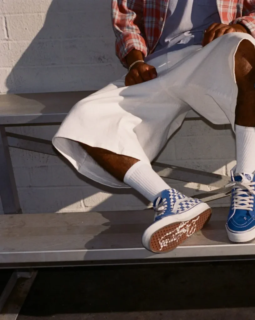 Person seated on bleachers wearing white shorts, plaid shirt and blue-and-white checkerboard sneakers