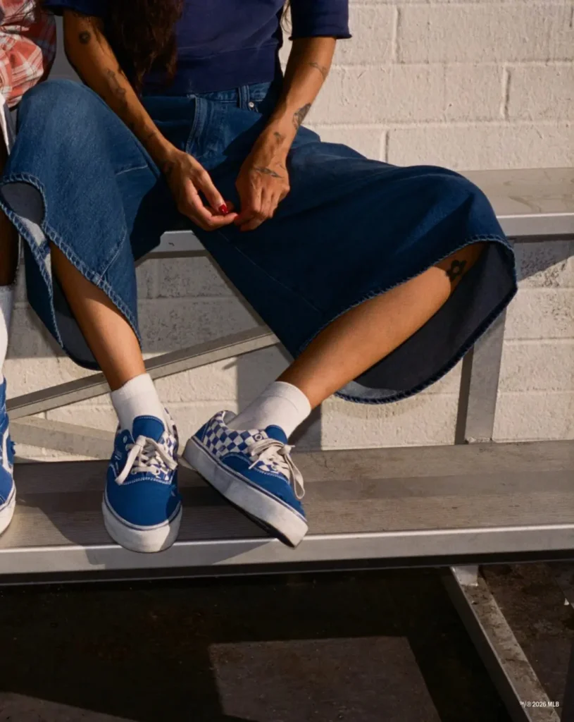 Seated person in a denim skirt wears blue-and-white sneakers on metal bleachers beside another person