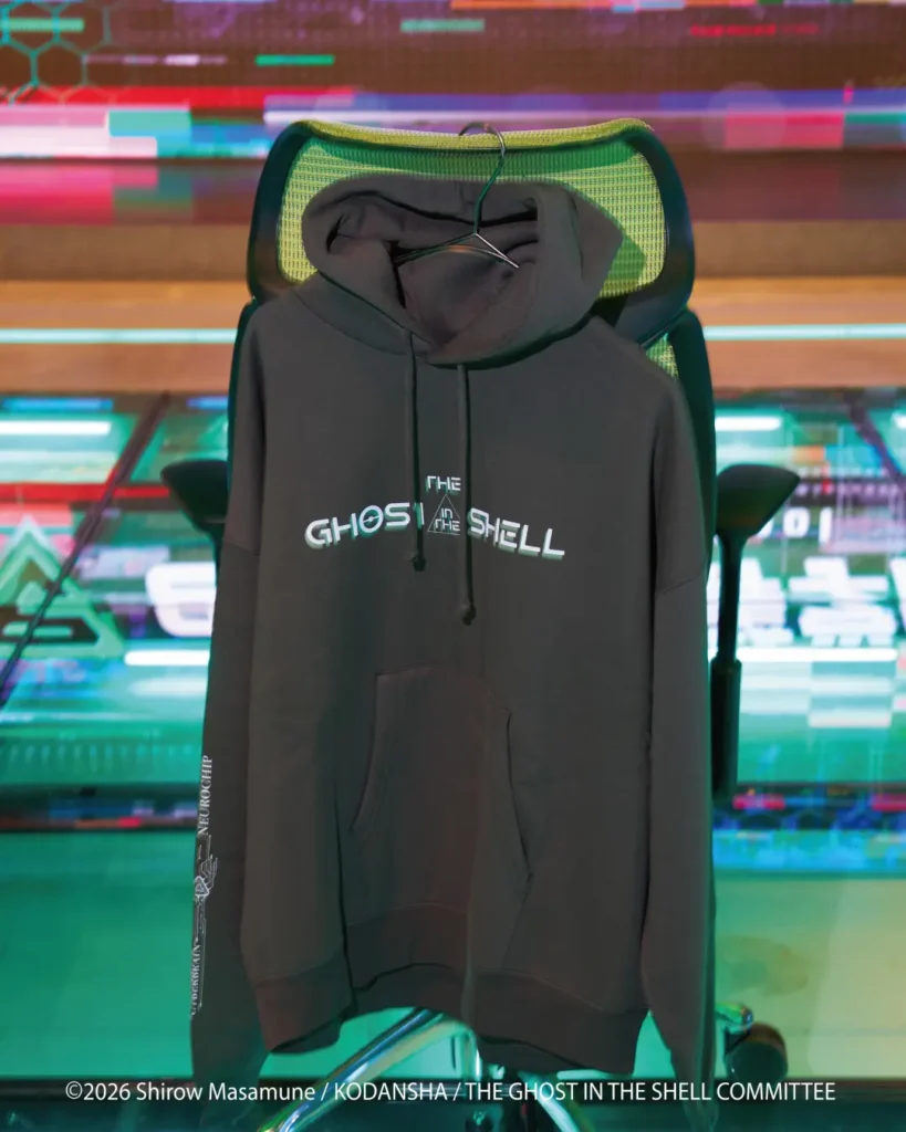 Black hooded sweatshirt hanging on a chair, with “THE GHOST IN THE SHELL” text and sleeve graphics, against a colorful