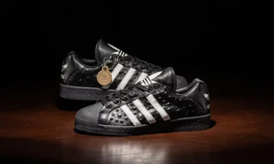 Two black leather Adidas sneakers with white stripes on a dark wooden surface, one overlapping the other