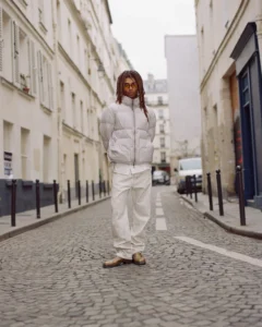Person in white puffer jacket and pants standing on a cobblestone street between urban buildings