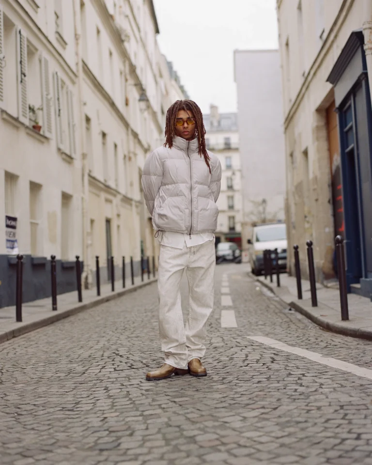 Person in white puffer jacket and pants standing on a cobblestone street between urban buildings
