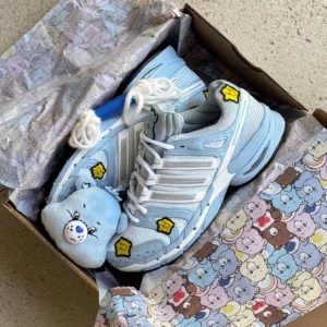Light blue and white sneakers with yellow cartoon patches and a matching blue plush toy in a patterned shoebox