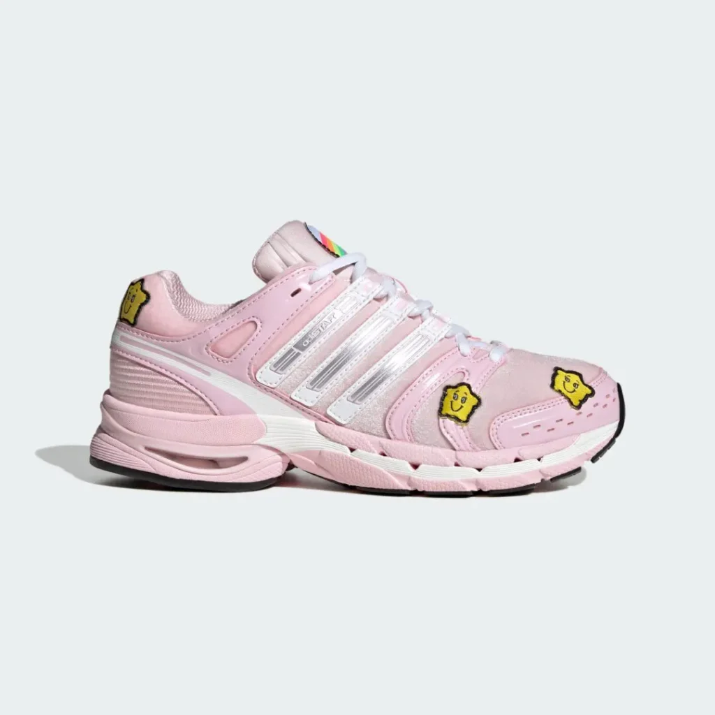 Pink Adidas sneaker with white stripes, yellow cartoon patches, rainbow tongue detail, and black-and-white sole on a pale