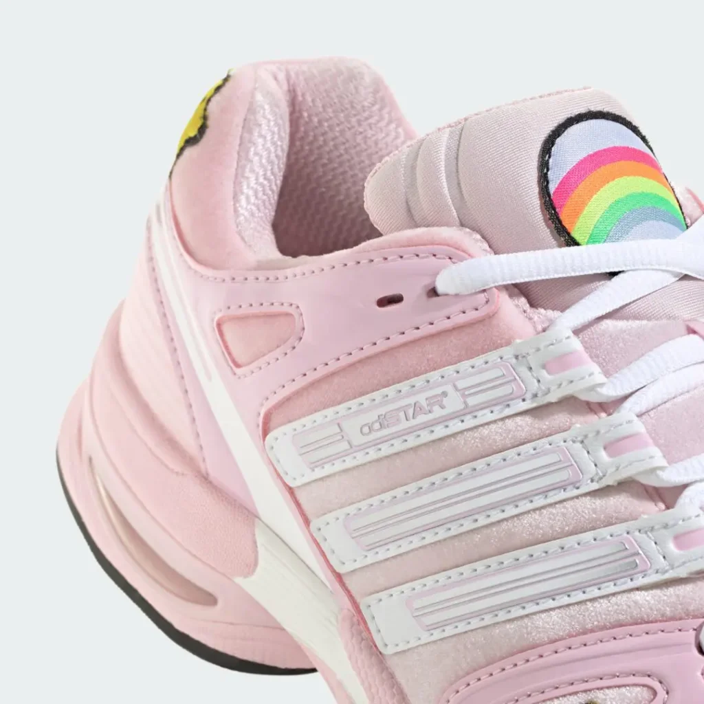 Close-up of a pink and white sneaker with layered straps, white laces, and a rainbow-striped tongue label against a pale