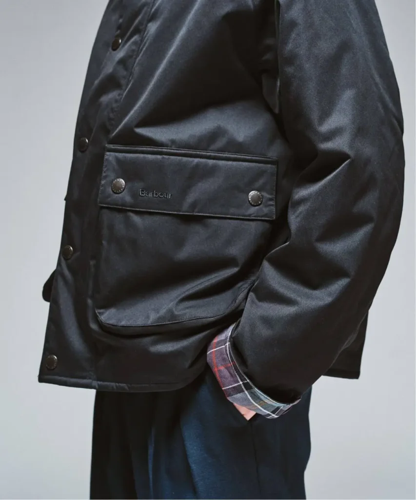 Close-up of a person wearing a black Barbour jacket with flap pockets, snap buttons and plaid cuff lining