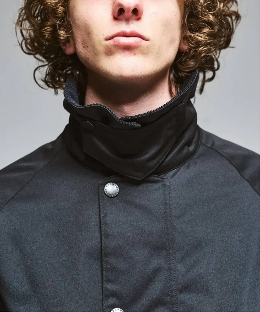Close-up portrait of a curly-haired person wearing a dark charcoal jacket with a high black collar and metal buttons against