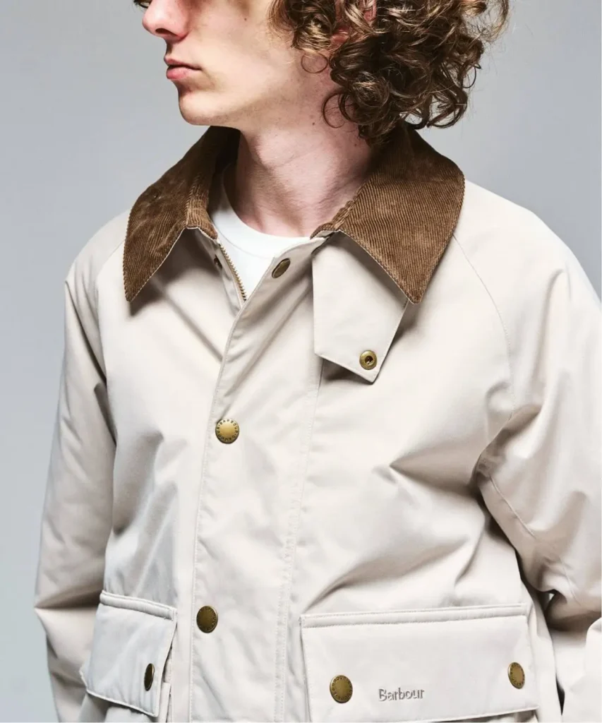 Cropped person in a cream Barbour jacket with a brown corduroy collar and multiple brass snap buttons against a gray