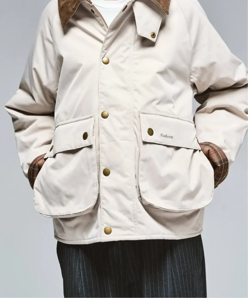 Close-up of a person wearing a cream Barbour jacket with corduroy collar, brass snaps, flap pockets, and plaid lining