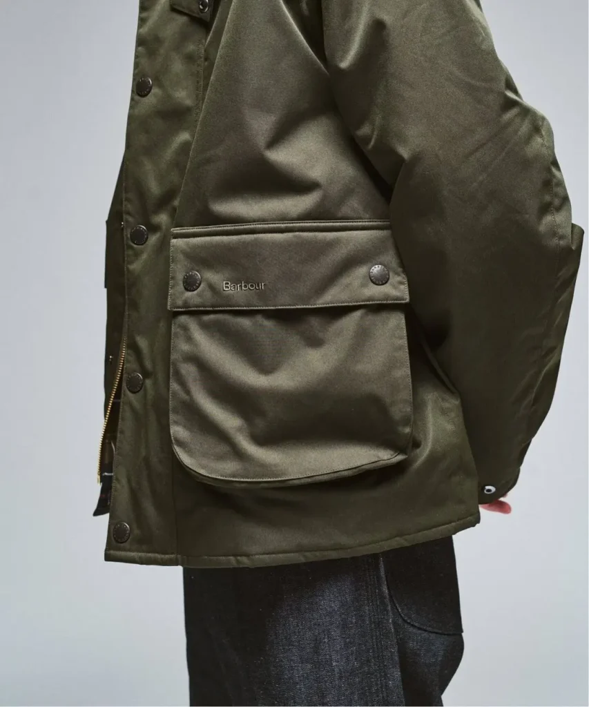 Close-up of a person wearing an olive waxed jacket with a Barbour-labeled flap pocket and black trousers against a pale
