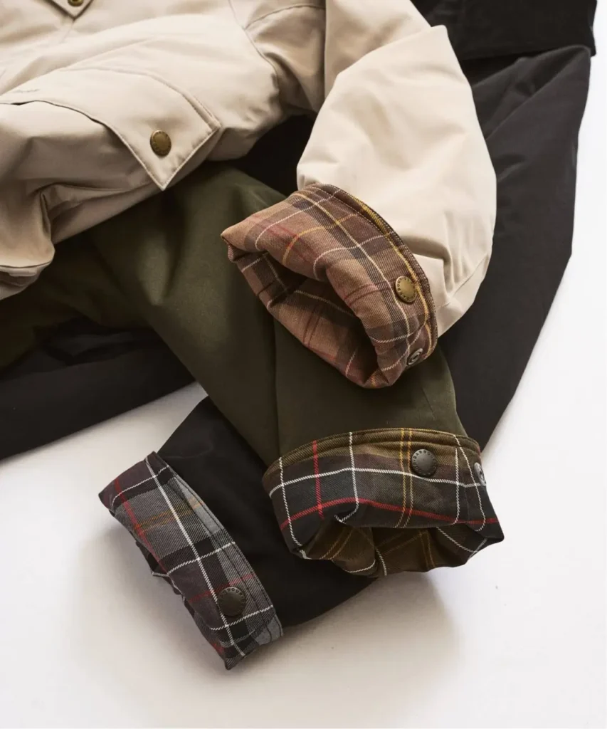 Close-up of a cream jacket with brown plaid cuffs layered over olive trousers and black fabric on a white surface