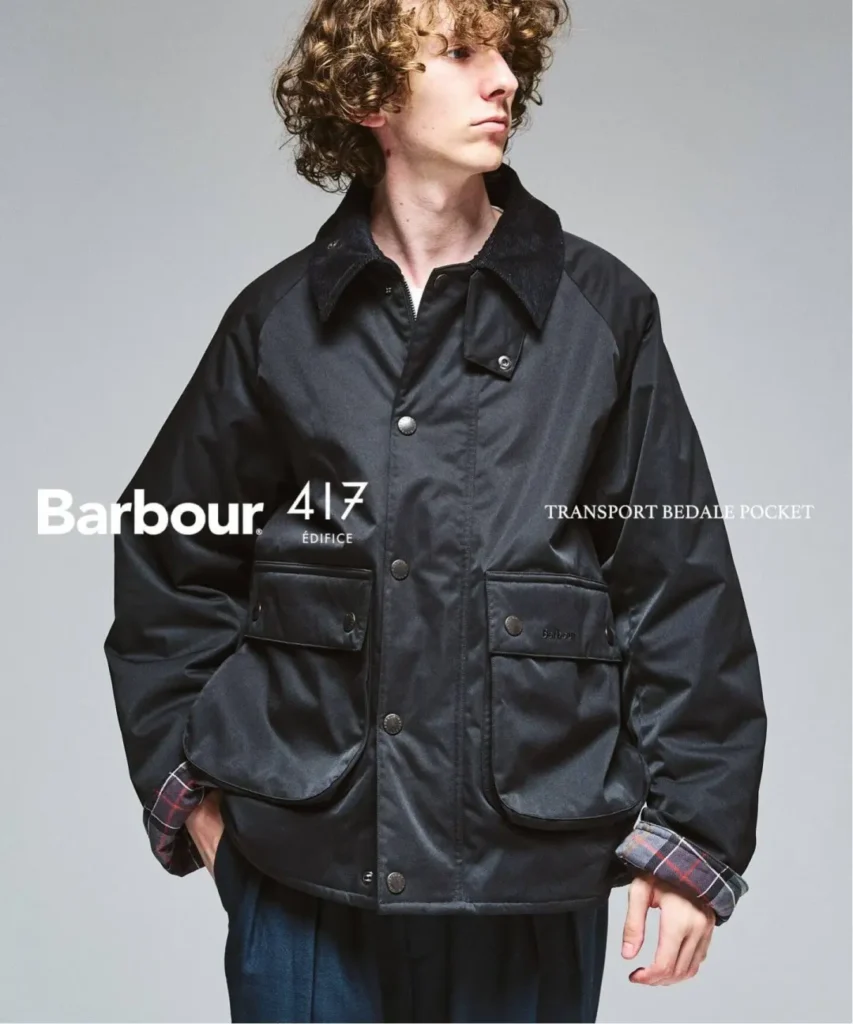 Curly-haired model in a black waxed jacket with plaid cuffs and multiple flap pockets, posed against a gray backdrop