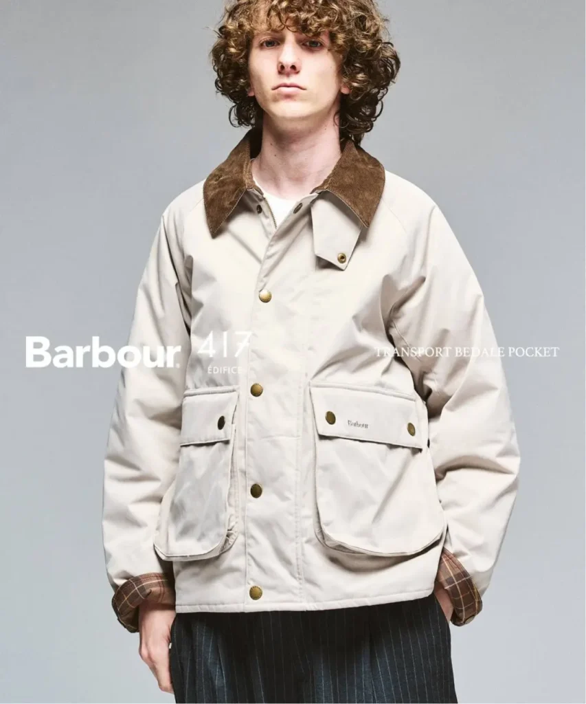 Curly-haired model wearing a cream Barbour jacket with a brown corduroy collar and large flap pockets against a gray