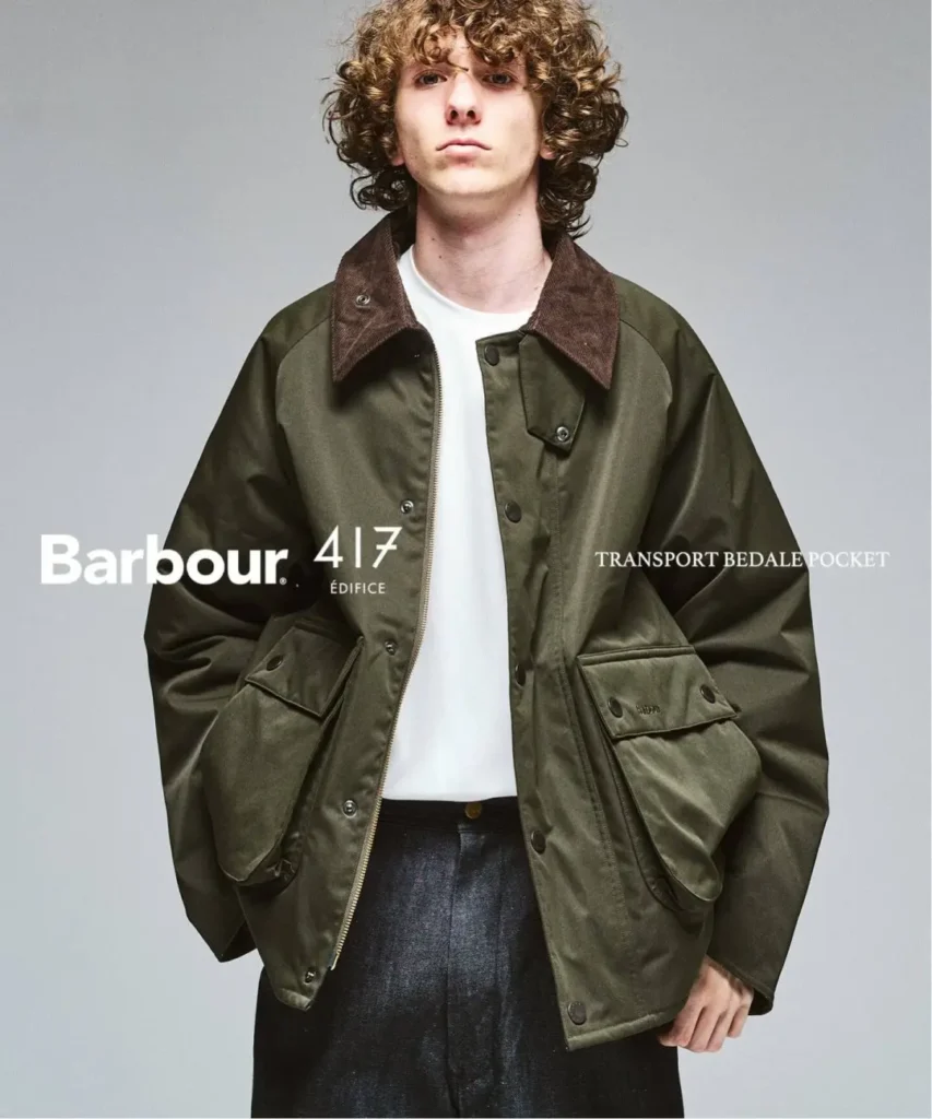 Curly-haired model wears an olive waxed jacket with brown corduroy collar and large flap pockets against a gray backdrop
